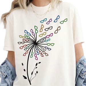 Könnte beinhalten: Ein elfenbeinfarbenes Kurzarm-T-Shirt mit einer schwarzen Grafik eines Löwenzahnstiels. Der Löwenzahnkopf besteht aus zahlreichen kleinen, bunten Brillengestellen in Blau, Grün, Rot, Gelb, Lila und Braun, die wie vom Wind verweht wirken.