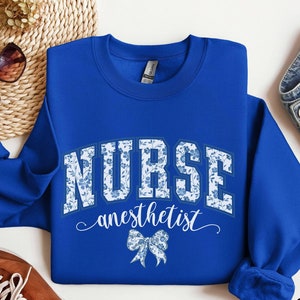 May include: Royal blue sweatshirt featuring the word "NURSE" in a floral pattern across the chest. The word "anesthetist" is written in cursive below, with a floral bow graphic. The sweatshirt is displayed on a woven surface.