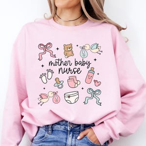 Könnte beinhalten: Ein hellrosa Sweatshirt mit dem Text "mother baby nurse" und babybezogenen Illustrationen, darunter ein Teddybär, ein Schnuller, eine Windel und eine Babyflasche.