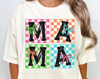 Op de afbeelding: Een wit T-shirt met een afbeelding van het woord "MAMA" in zwarte letters met kleurrijke strikken op elke letter. De letters staan op een geruite achtergrond met afwisselende kleuren roze, oranje, groen en blauw.