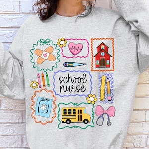 May include: A light gray sweatshirt featuring colorful school-themed graphics. The design includes a bandage, heart, schoolhouse, crayons, thermometer, ruler, pencil, ice pack, school bus, stethoscope, and the words "school nurse".