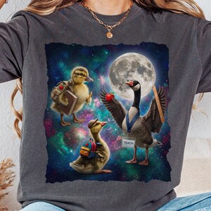 Pode incluir: Uma t-shirt cinzento-carvão com um gráfico de dois patinhos e um ganso no espaço. Os patinhos usam mochilas e carregam livros, enquanto o ganso segura uma maçã e uma régua, usando uma etiqueta com a palavra TEACH sob a lua cheia.