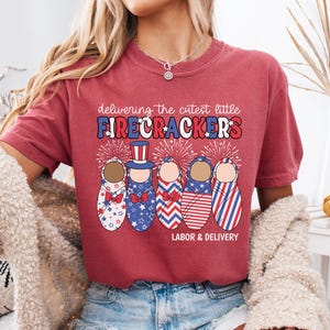 Könnte beinhalten: Rotes T-Shirt mit einer Grafik von fünf Babyfiguren, die in rot-weiß-blauen patriotischen Outfits gekleidet sind. Der Text "delivering the cutest little FIRECRACKERS LABOR & DELIVERY" ist auf das T-Shirt gedruckt.
