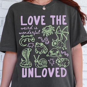 May include: A charcoal grey short-sleeve t-shirt featuring the light purple text LOVE THE UNLOVED and weird is wonderful. The graphic includes light green line art illustrations of a spider, bat, lizard, snake, snail, frog, blobfish, and mouse.