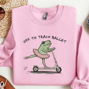 May include: A light pink crewneck sweatshirt featuring a graphic of a green frog wearing a pink tutu and ballet slippers while riding a kick scooter. The text above the frog reads "OFF TO TEACH BALLET" in black capital letters.