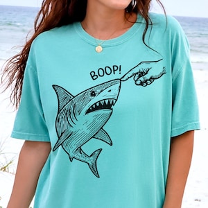 Pu&ograve; includere: T-shirt verde acqua con grafica nera di uno squalo a cui viene toccato il naso con un dito, con la scritta "BOOP!" sopra. La maglietta &egrave; a tinta unita e ha maniche corte.