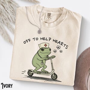 Puede incluir: Sudadera de color marfil con el texto "OFF TO HELP HEARTS" y una rana de dibujos animados con gorro de enfermera y montada en un patinete. Un collar plateado con un colgante está encima de la camisa.