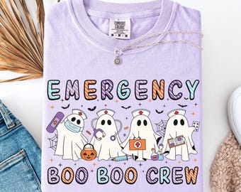 May include: A light purple Comfort Colors t-shirt with the words "EMERGENCY BOO BOO CREW" in colorful letters. The shirt features cartoon ghost characters dressed as nurses, holding medical supplies and a pumpkin. A silver necklace with a sun pendant is draped across the shirt.