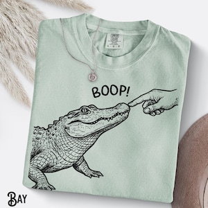May include: A pale green t-shirt featuring a black and white alligator illustration being touched on the nose. The word "BOOP!" is printed above the alligator. A silver necklace with a pendant is draped on the shirt.