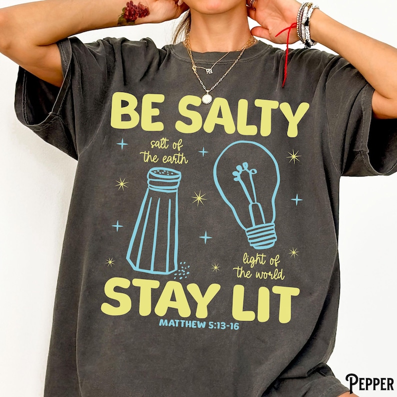 Puede incluir: Camiseta gris oscuro con las palabras "BE SALTY STAY LIT" en amarillo, con ilustraciones azules de un salero y una bombilla. Tambi&eacute;n se incluye el texto "sal de la tierra" y "luz del mundo". El vers&iacute;culo b&iacute;blico Mateo 5:13-16 est&aacute; impreso en la parte inferior.