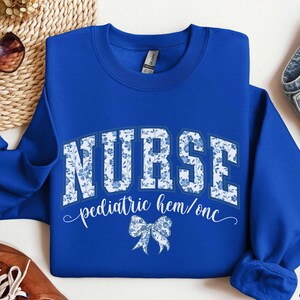 May include: Royal blue sweatshirt with the word "NURSE" in white block letters filled with a blue floral pattern. Below the word "NURSE" is the text "pediatric hem/onc" in white script, and a blue bow graphic.