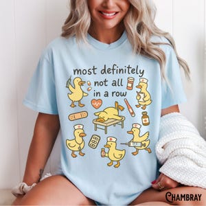 Puede incluir: Camiseta azul claro con un dise&ntilde;o divertido de patos de dibujos animados en situaciones m&eacute;dicas. El texto "most definitely not all in a row" se muestra encima de las ilustraciones. Los patos est&aacute;n representados con herramientas m&eacute;dicas.
