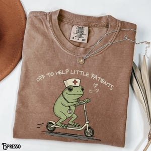 May include: Espresso-colored t-shirt with the phrase "OFF TO HELP LITTLE PATIENTS" and a cartoon frog wearing a nurse's hat and riding a scooter. The shirt has a Comfort Colors tag and a silver necklace.