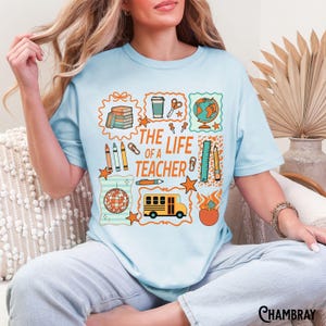May include: Light blue t-shirt with a colorful graphic design. The design includes the text "THE LIFE OF A TEACHER" with illustrations of school supplies, a globe, and a school bus. The shirt is a casual, everyday wear item.