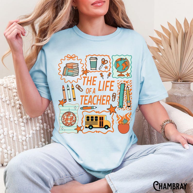 May include: Light blue t-shirt with a colorful graphic design. The design includes the text "THE LIFE OF A TEACHER" with illustrations of school supplies, a globe, and a school bus. The shirt is a casual, everyday wear item.