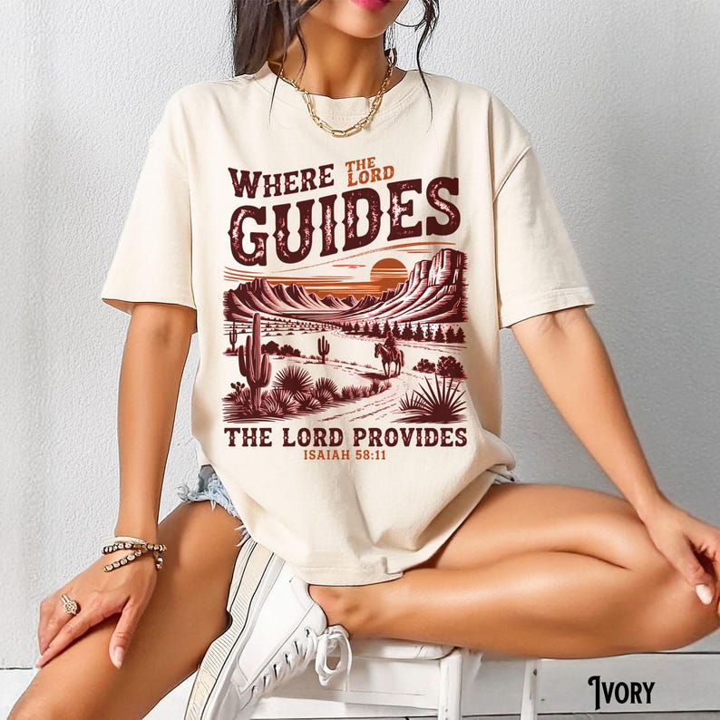 May include: A beige t-shirt with a graphic print of a sunset landscape with a cowboy riding a horse. The text "Where the Lord Guides The Lord Provides Isaiah 58:11" is printed on the shirt.