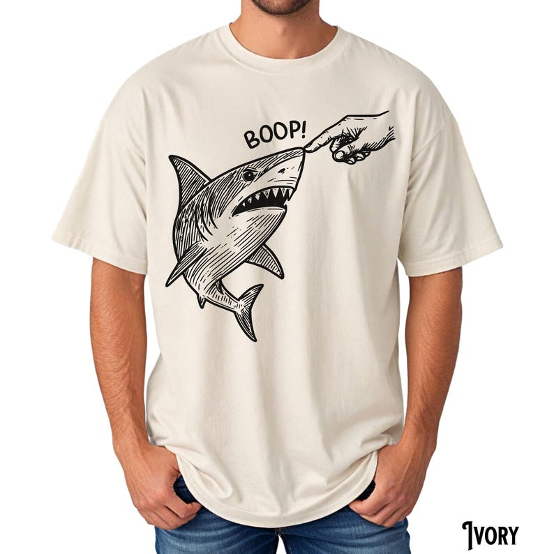 Pu&ograve; includere: T-shirt color avorio con un'illustrazione in bianco e nero di uno squalo a cui viene toccato il naso. La parola "BOOP!" &egrave; sopra lo squalo. La maglietta &egrave; realizzata in un materiale morbido.