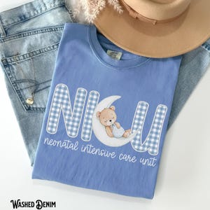 Op de afbeelding: Lichtblauw T-shirt met de letters "NICU" in een blauw-wit ruitpatroon. Een teddybeer slaapt op een maan. De woorden "neonatal intensive care unit" staan eronder.