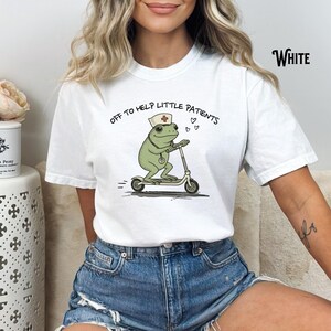 May include: White t-shirt featuring a cartoon frog wearing a nurse's hat and riding a scooter. The frog has a stethoscope and the text "OFF TO HELP LITTLE PATIENTS" is printed above the frog. The word "White" is printed on the upper right.