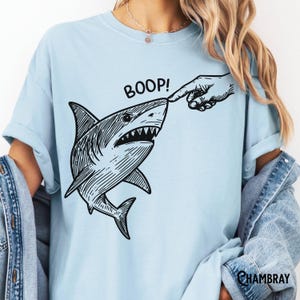 Pu&ograve; includere: T-shirt azzurra con un'illustrazione in bianco e nero di uno squalo toccato sul naso da un dito. La parola "BOOP!" &egrave; stampata sopra lo squalo. La maglietta ha maniche corte e una vestibilit&agrave; ampia.
