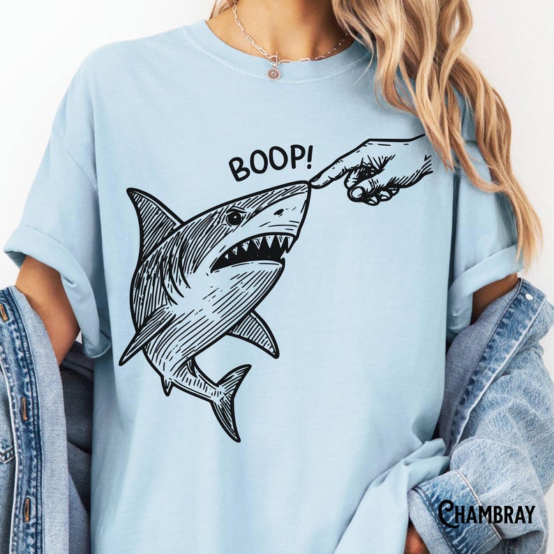 Pu&ograve; includere: T-shirt azzurra con un'illustrazione in bianco e nero di uno squalo toccato sul naso da un dito. La parola "BOOP!" &egrave; stampata sopra lo squalo. La maglietta ha maniche corte e una vestibilit&agrave; ampia.