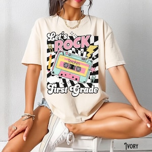 May include: Ivory-colored t-shirt with a retro graphic. The design features the words "Let's Rock First Grade" with a cassette tape illustration in pastel colors. A pencil, lightning bolt, and checkerboard pattern complete the design.