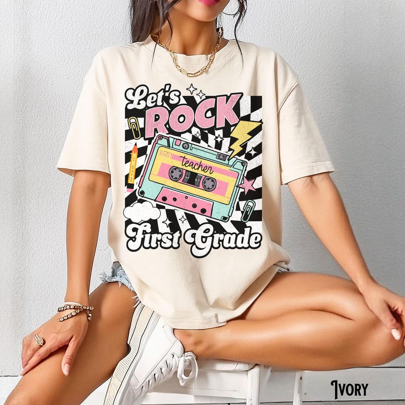 May include: Ivory-colored t-shirt with a retro graphic. The design features the words "Let's Rock First Grade" with a cassette tape illustration in pastel colors. A pencil, lightning bolt, and checkerboard pattern complete the design.