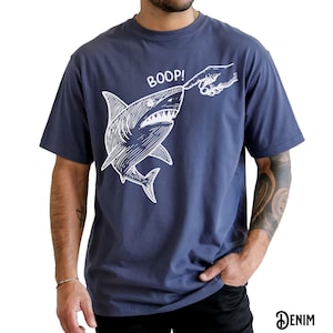 Pu&ograve; includere: T-shirt blu navy con grafica bianca di uno squalo che viene toccato sul naso da un dito, con la scritta "BOOP!" sopra. La parola "DENIM" &egrave; stampata in basso a destra.
