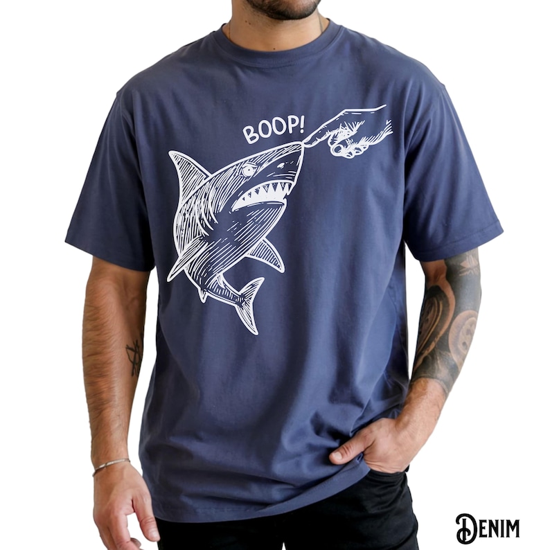 Pu&ograve; includere: T-shirt blu navy con grafica bianca di uno squalo che viene toccato sul naso da un dito, con la scritta "BOOP!" sopra. La parola "DENIM" &egrave; stampata in basso a destra.