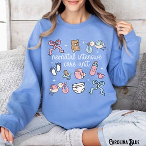 Op de afbeelding: Een lichtblauw sweatshirt met de tekst "neonatal intensive care unit" en verschillende baby-thema illustraties, waaronder een teddybeer, een ooievaar, een speen en een paar sokken.