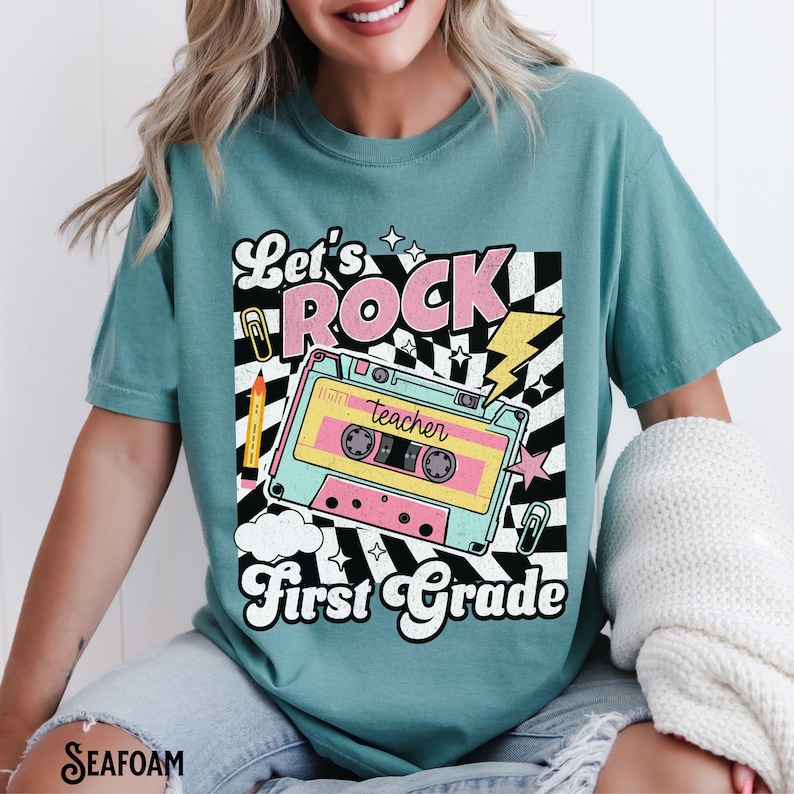 May include: Seafoam green t-shirt with a retro design. The graphic features the words "Let's Rock First Grade" with a cassette tape, lightning bolt, and other school-themed elements. The shirt is a comfortable, casual style.