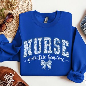 May include: Royal blue sweatshirt with the word "NURSE" in white block letters filled with a blue floral pattern. Below the word "NURSE" is the text "pediatric hem/onc" and a blue bow graphic. The sweatshirt is a crew neck style.