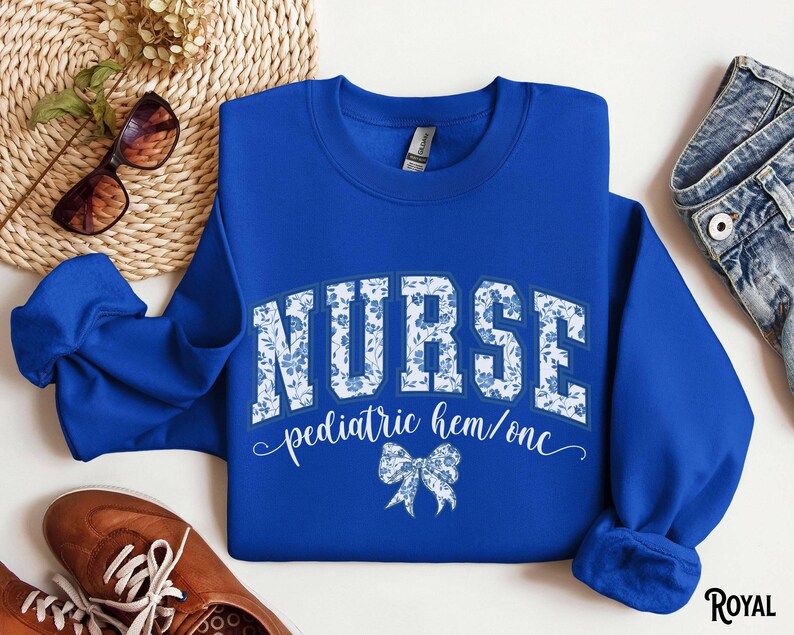 May include: Royal blue sweatshirt with the word "NURSE" in white block letters filled with a blue floral pattern. Below the word "NURSE" is the text "pediatric hem/onc" and a blue bow graphic. The sweatshirt is a crew neck style.