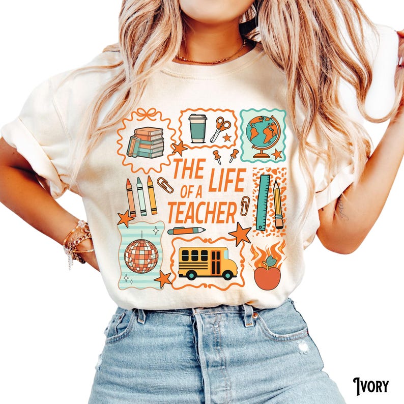 May include: Ivory-colored t-shirt with a colorful graphic that reads "The Life of a Teacher." The design includes school-related icons such as books, a globe, pencils, a school bus, and an apple. The shirt has short sleeves and a crew neck.