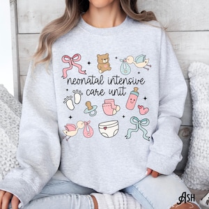 Op de afbeelding: Een grijs sweatshirt met de tekst "neonatal intensive care unit" en baby-thema illustraties, waaronder een teddybeer, een ooievaar, een speen, een fles en een luier.