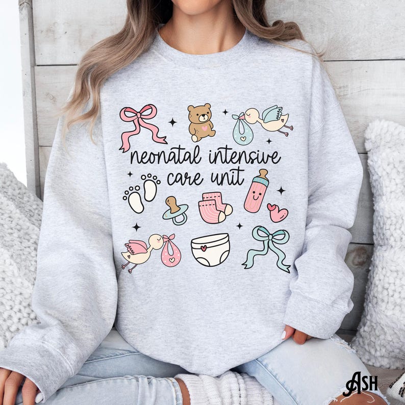 Op de afbeelding: Een grijs sweatshirt met de tekst "neonatal intensive care unit" en baby-thema illustraties, waaronder een teddybeer, een ooievaar, een speen, een fles en een luier.