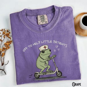 May include: A purple Comfort Colors t-shirt with the text "OFF TO HELP LITTLE PATIENTS" and a cartoon frog wearing a nurse's hat and riding a scooter. The shirt is a grape color.
