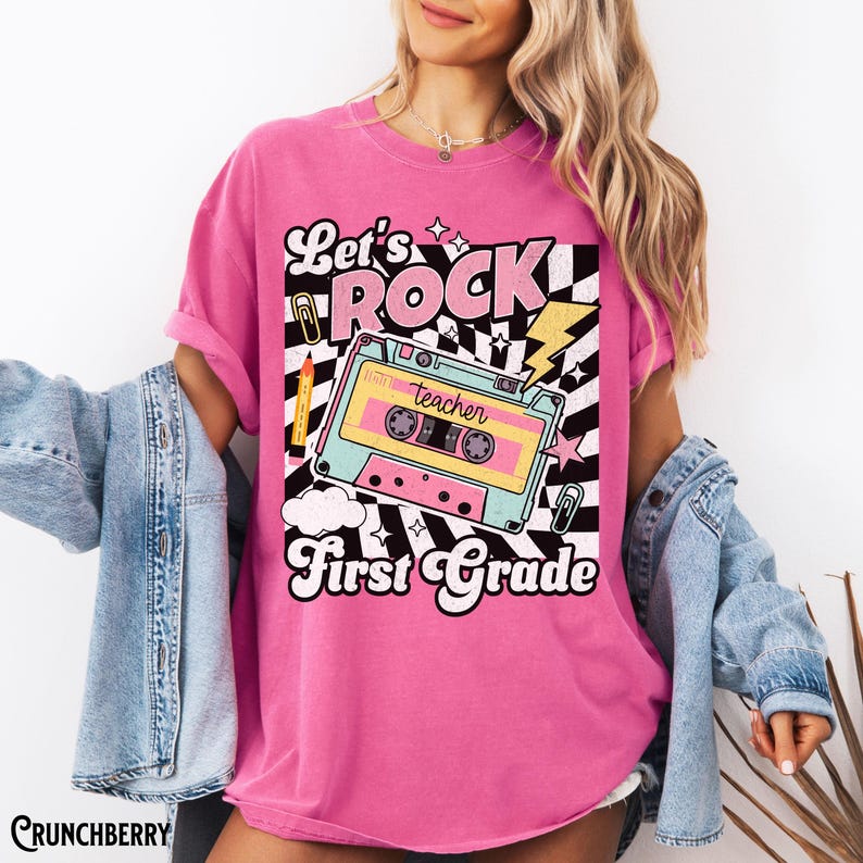 May include: A pink t-shirt with a graphic design featuring a retro cassette tape with the word "teacher" on it. The design includes the words "Let's ROCK First Grade" in a playful font, with stars, a lightning bolt, and a checkered background.