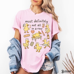Puede incluir: Camiseta rosa con la frase "most definitely not all in a row" e ilustraciones de patos amarillos con gorros de enfermera y material m&eacute;dico. La camiseta tiene un corte holgado.