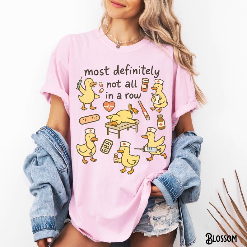 Puede incluir: Camiseta rosa con la frase "most definitely not all in a row" e ilustraciones de patos amarillos con gorros de enfermera y material m&eacute;dico. La camiseta tiene un corte holgado.