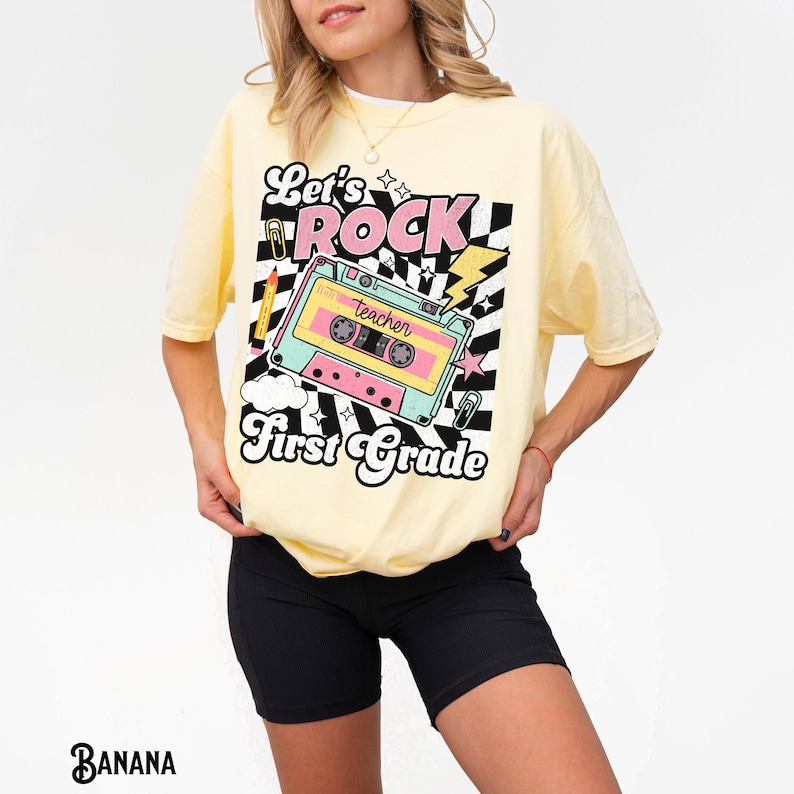 May include: A light yellow t-shirt with a retro graphic design. The design features the words "Let's ROCK First Grade" with a cassette tape illustration in pink, teal, and yellow. The shirt is a comfortable, oversized fit.