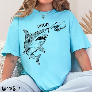Pu&ograve; includere: T-shirt blu laguna con grafica in bianco e nero di uno squalo toccato sul naso da un dito. La parola "BOOP!" &egrave; sopra lo squalo. Le parole "Lagoon Blue" sono in fondo alla maglietta.