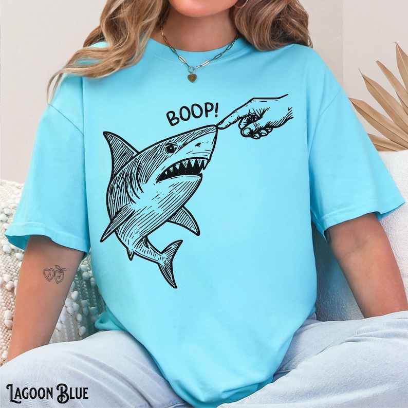 Pu&ograve; includere: T-shirt blu laguna con grafica in bianco e nero di uno squalo toccato sul naso da un dito. La parola "BOOP!" &egrave; sopra lo squalo. Le parole "Lagoon Blue" sono in fondo alla maglietta.
