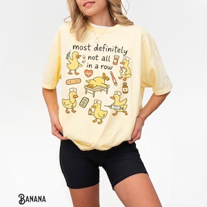 Puede incluir: Camiseta amarillo claro con el texto "most definitely not all in a row" y patos de dibujos animados vestidos con atuendos m&eacute;dicos. Los patos sostienen suministros m&eacute;dicos. La camiseta es de corte holgado.