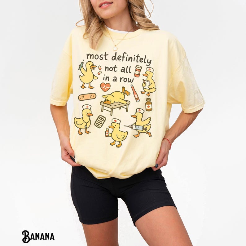 Puede incluir: Camiseta amarillo claro con el texto "most definitely not all in a row" y patos de dibujos animados vestidos con atuendos m&eacute;dicos. Los patos sostienen suministros m&eacute;dicos. La camiseta es de corte holgado.