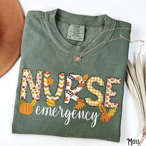 May include: A moss green t-shirt with the words "NURSE emergency" in a fall-themed design. The letters are filled with autumn leaves, pumpkins, and plaid patterns. A gold necklace with a pendant rests on the shirt.