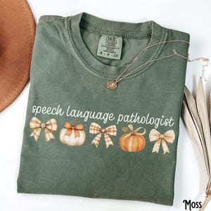 Può includere: Una maglietta verde muschio con le parole "speech language pathologist" stampate in bianco. La maglietta presenta un disegno di zucche e fiocchi. Una collana d'oro con un ciondolo è drappeggiata sulla maglietta.