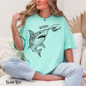 Pu&ograve; includere: T-shirt verde acqua con un'illustrazione in bianco e nero di uno squalo che viene toccato sul naso da un dito. La parola "BOOP!" &egrave; sopra lo squalo. Il marchio "ISLAND REEF" &egrave; stampato in basso.