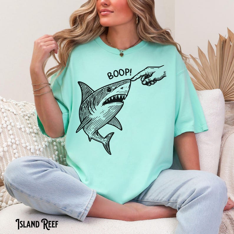 Pu&ograve; includere: T-shirt verde acqua con un'illustrazione in bianco e nero di uno squalo che viene toccato sul naso da un dito. La parola "BOOP!" &egrave; sopra lo squalo. Il marchio "ISLAND REEF" &egrave; stampato in basso.