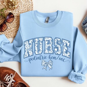 May include: Light blue sweatshirt with the word "NURSE" in large, floral-patterned letters. Below, it reads "pediatric hem/onc" with a bow graphic. The sweatshirt is surrounded by accessories, including sunglasses and shoes.